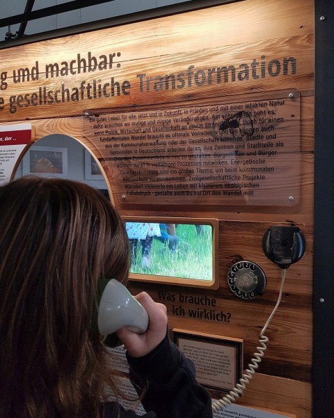 Person hält einen Telefonhörer ans Ohr und schaut dabei auf kleinen Bildschirm, im Hintergrund steht "machbar gesellschaftliche Transformation"